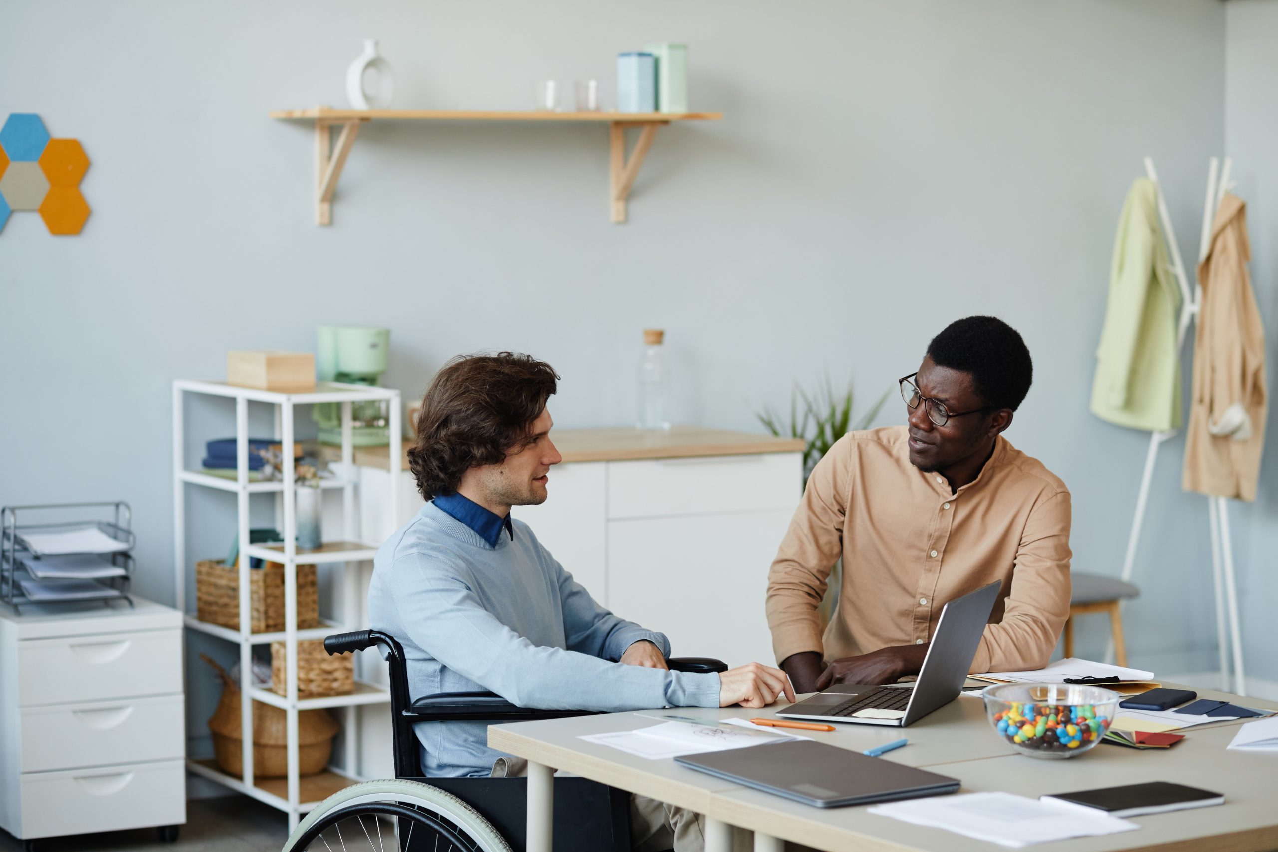 Two men in a campus office discuss accessibility planning. One uses a wheelchair, while the other works on a laptop with documents and reports nearby. The setting reflects a higher ed environment focused on managing student accommodations and support.