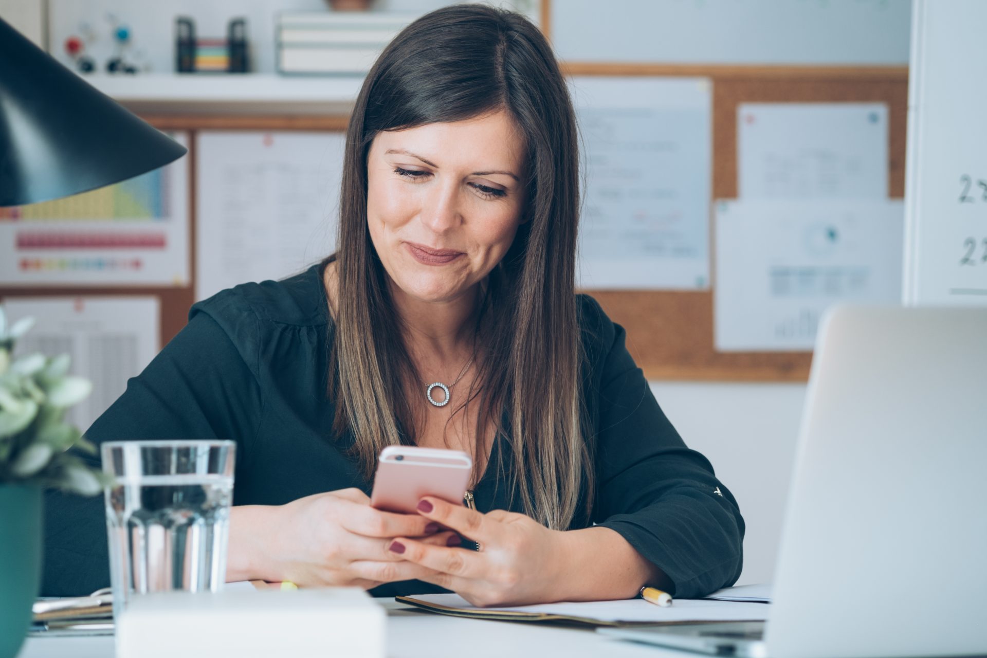 A woman smiles while reading a message on her phone at her desk, representing how the message center helps streamline communication.