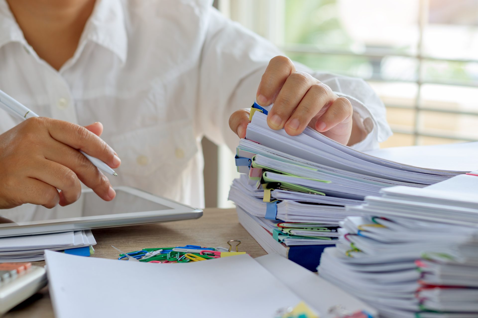 A person reviewing large stacks of paperwork beside a tablet, representing outdated accommodation systems that rely on manual processes.