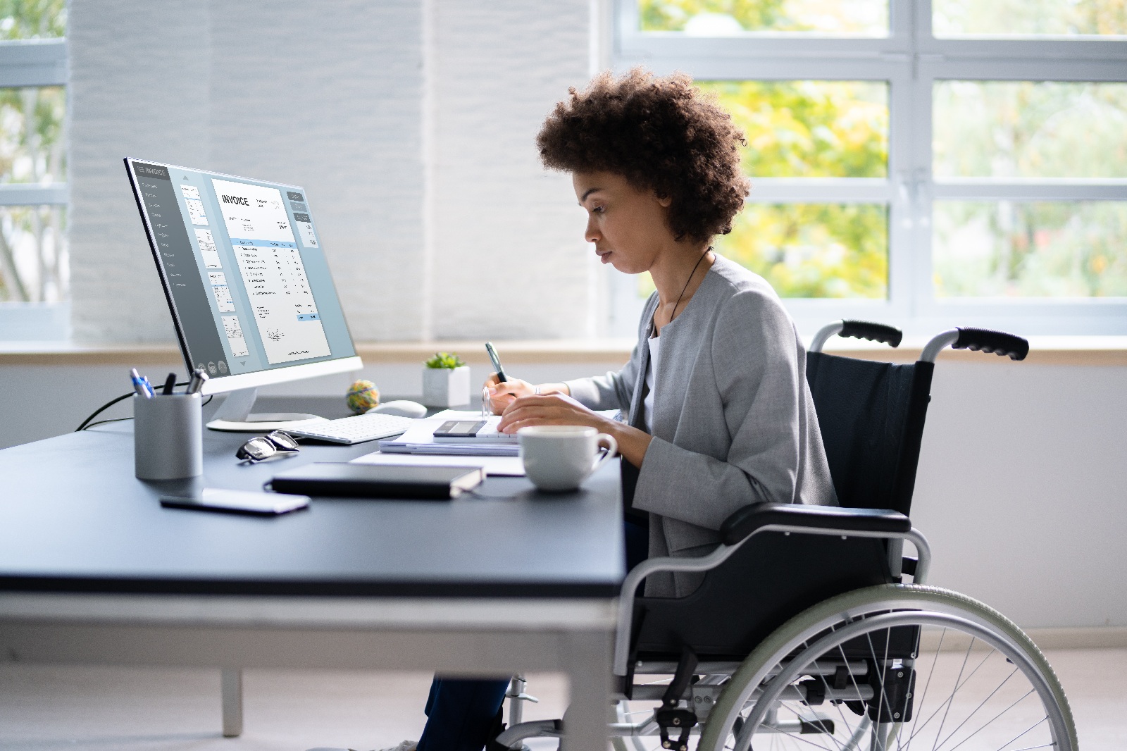 Person in a wheelchair working at a desk with a computer and paperwork in an office.