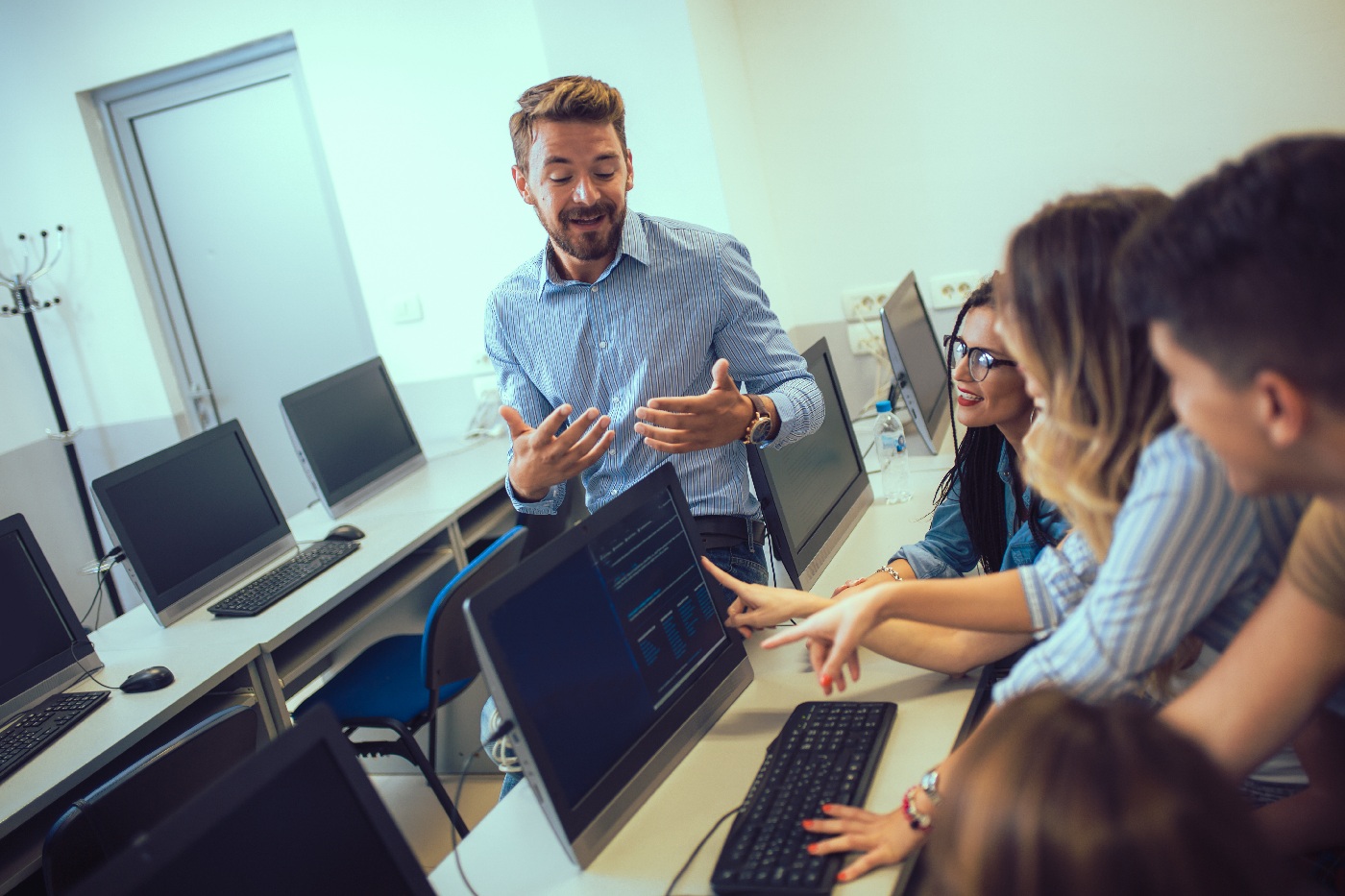 Teacher stands beside college students working at desktop computers in a classroom.
